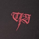 Detailfotografie einer rotfarbigen Stickerei auf schwarzer Thor-Steinar-Kleidung mit einem T und S in altertümlicher Schrift sowie einem Blitz-Symbol.