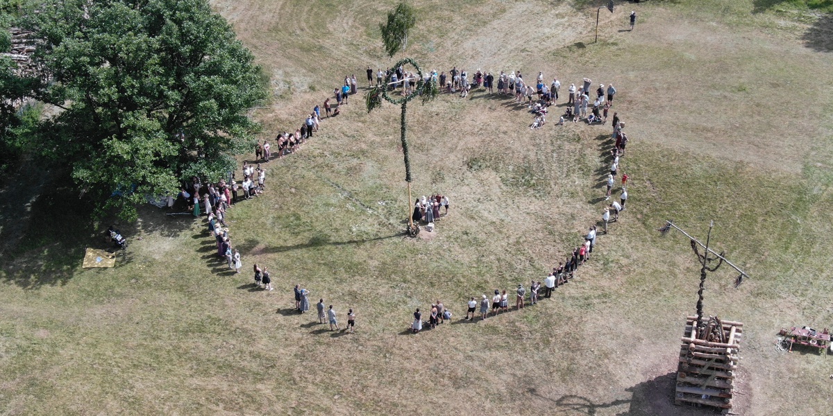 Eine Fotoaufnahme aus der Vogelperspektive auf eine Gruppe volkstümlich gekleideter Menschen, die auf einer Wiese in einem Kreis um eine Säule stehen, die ein Keltenkreuz zeigt.