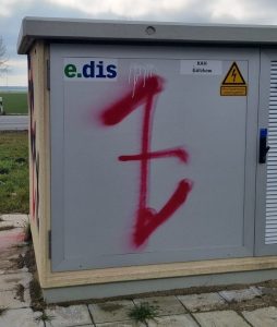 Foto eines roten Graffiti einer aufrechten, gespiegelte Gibor-Rune bzw. Wolfsangel auf einem Stromkasten