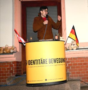 Foto von Martin Sellner an einem Rednerpult der Identitären Bewegung mit Deutschland- und Österreichfahne vor einem Hauseingang.