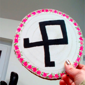 Auf eine Baumscheibe gemalte Odal-Rune mit rosa Blütenkranz.