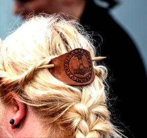 Foto einer Haarnadel mit einem Stück Leder, auf welchem ein Thorshammer eingebrannt ist, in blondem, geflochtenen Haar.